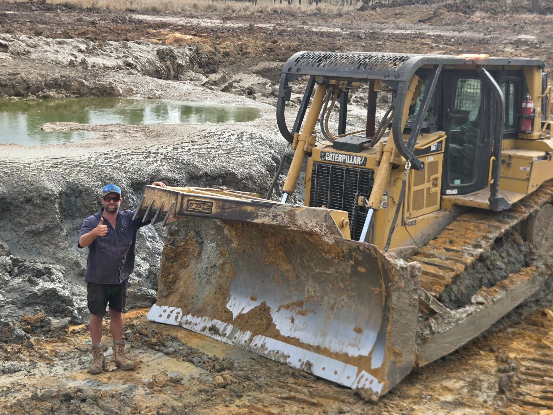 Tyrone giving a thumbs up next to the blade of a CAT D6T dozer beside a freshly cut dam