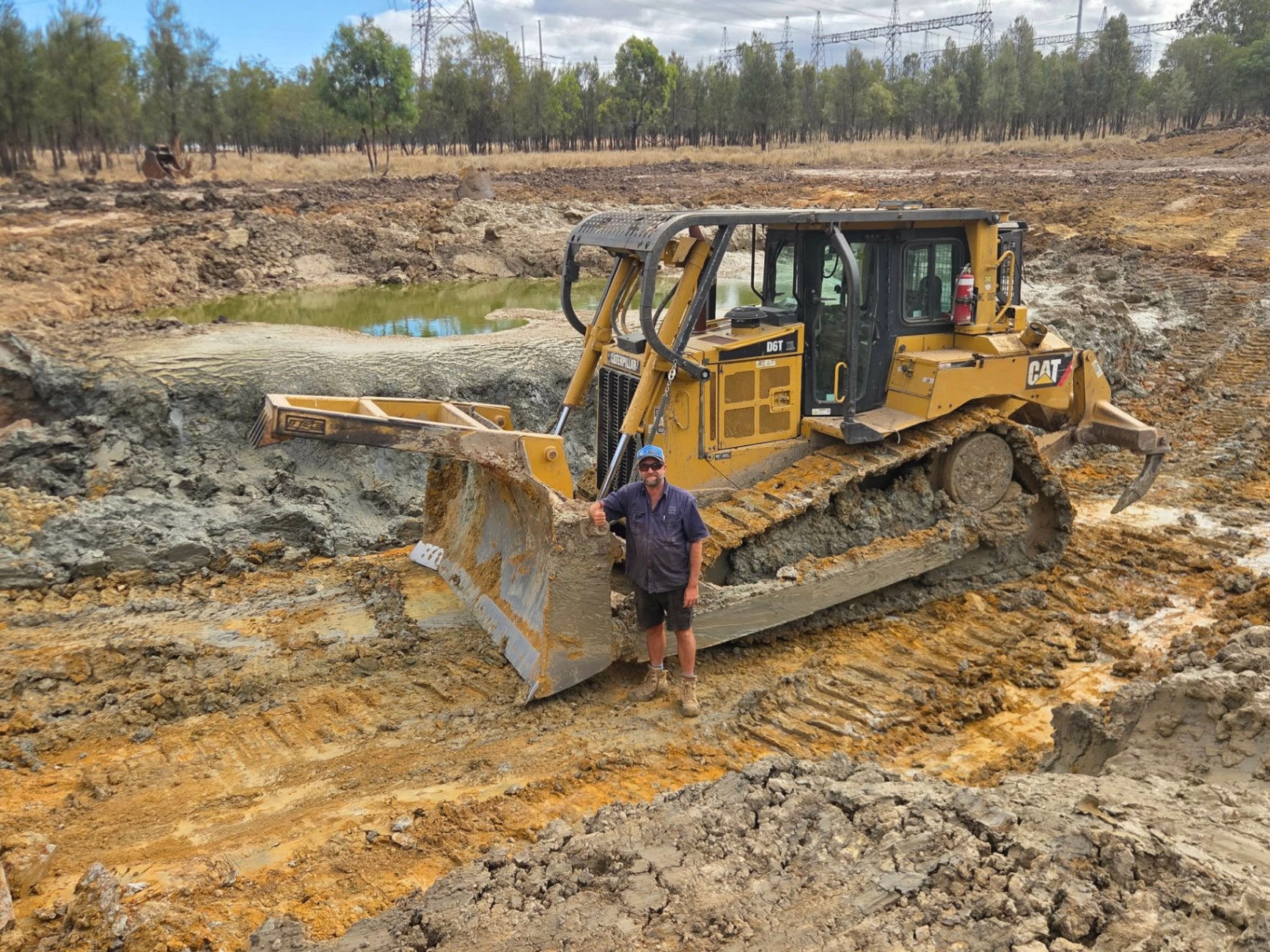 Tyrone standing in front of a CAT D6T dozer beside a partly filled dam under powerlines