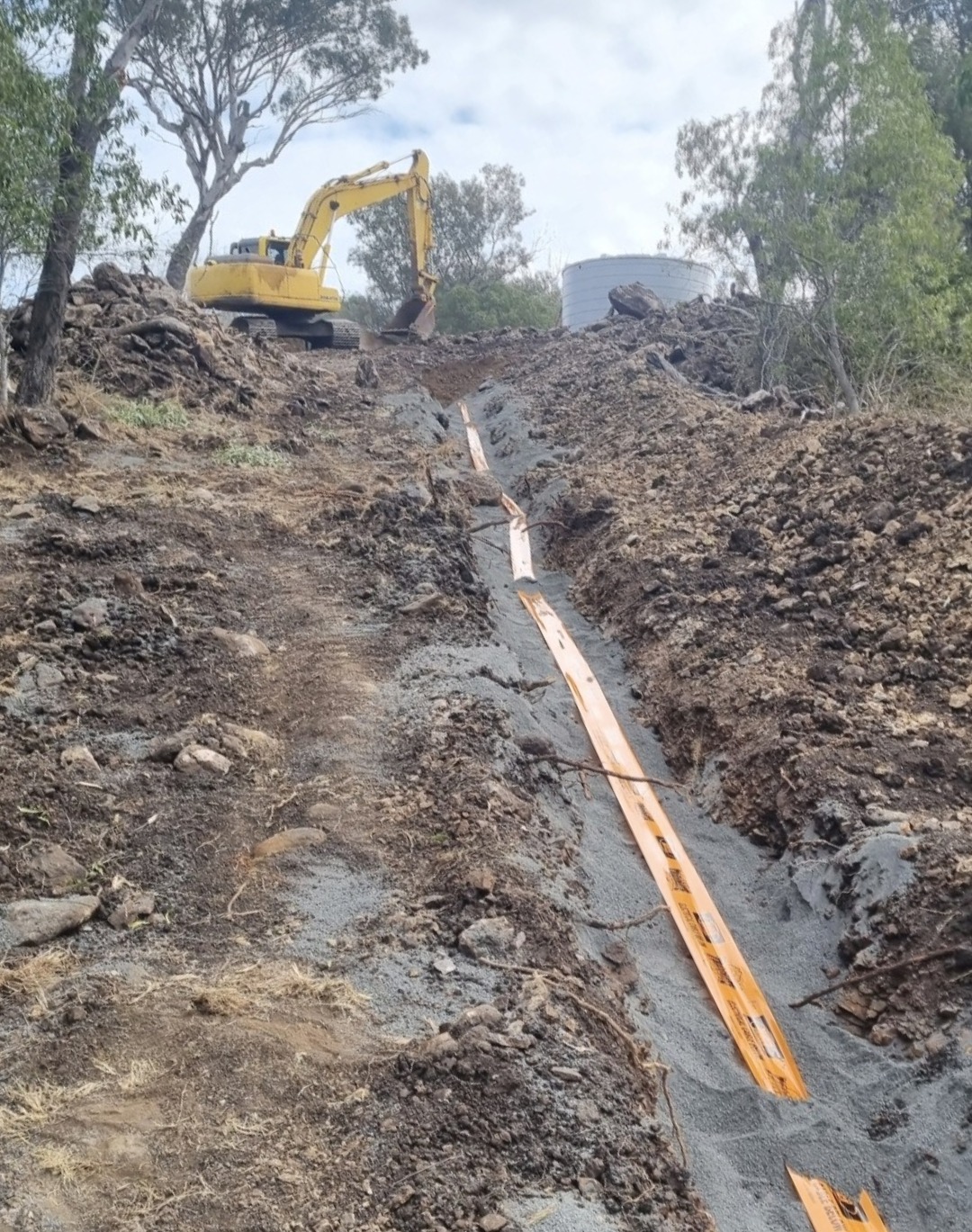Excavator on a hilltop beside a freshly cut trench with orange poly pipe running down to a water tank